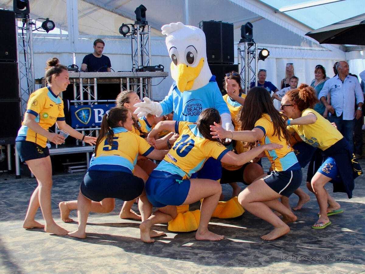 Group of Italian Players in Rugby Uniforms Dancing With Mascot Gullfy on a Beach